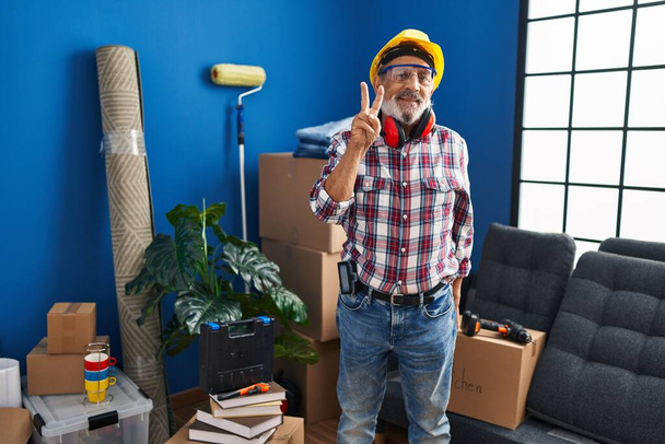 Cheerful senior man in hardhat and safety glasses, proudly showing the 'two' sign with fingers while joyously pointing up at his new home - Photo, Image