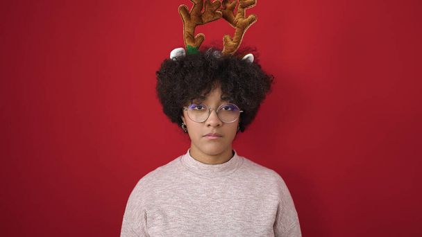 Νεαρή Αφροαμερικανή γυναίκα που φοράει χριστουγεννιάτικα αυτιά ταράνδου με σοβαρή έκφραση πάνω από απομονωμένο κόκκινο φόντο - Φωτογραφία, εικόνα