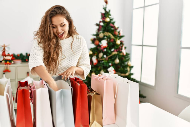 Νεαρή όμορφη ισπανόφωνη γυναίκα ψάχνει τσάντες ψώνια στέκεται δίπλα στο χριστουγεννιάτικο δέντρο στο σπίτι - Φωτογραφία, εικόνα