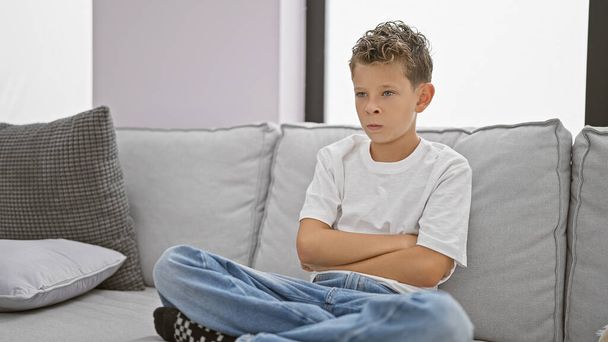 Imádnivaló szőke fiú ül egy hangulatos nappaliban, karok keresztben egy feldúlt kifejezés, sugárzó komoly vibrálást a kényelmes kanapén otthon. - Fotó, kép