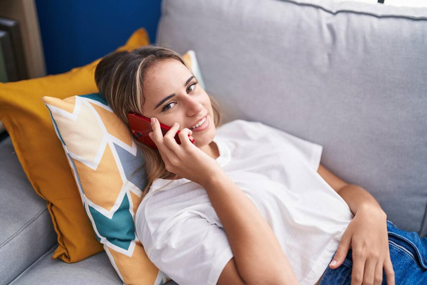 Jeune femme blonde parlant sur le smartphone couché sur le canapé à la maison - Photo, image