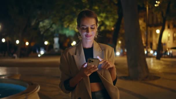 Menina feliz navegando celular à noite ao ar livre. Mulher de negócios sorrindo desfrutando de comunicação online verificando as mídias sociais na rua da cidade. Mulher elegante tocando celular compras em luzes da noite - Filmagem, Vídeo