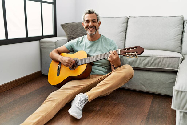 Grauhaariger Mann mittleren Alters sitzt zu Hause auf dem Boden und spielt klassische Gitarre - Foto, Bild