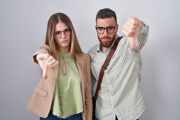 Young couple standing over white background looking unhappy and angry showing rejection and negative with thumbs down gesture. bad expression.  - Photo, Image
