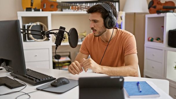 Homem hispânico com fones de ouvido fala em microfone em casa estúdio, retratando uma cena criativa ou podcasting. - Foto, Imagem