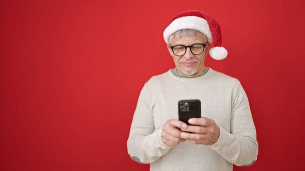 Μεσήλικας γκριζομάλλης άντρας που φοράει χριστουγεννιάτικο καπέλο χρησιμοποιώντας smartphone σε απομονωμένο κόκκινο φόντο - Φωτογραφία, εικόνα