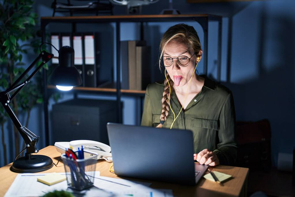 Young blonde woman working at the office at night sticking tongue out happy with funny expression. emotion concept.  - Foto, Imagem