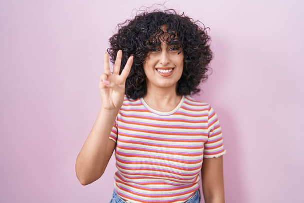 若い中東の女性がピンクの背景の上に立って自信と幸せに微笑んでいる間指番号3を指で指で指を指さす.  - 写真・画像