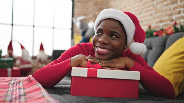Afroamerikanerin mit Weihnachtsmütze stützt sich auf Geschenk zu Hause - Foto, Bild