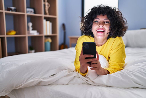 Jonge mooie Spaanse vrouw met behulp van smartphone liggend op bed in de slaapkamer - Foto, afbeelding