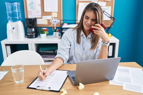Młoda piękna latynoska kobieta biznes pracownik mówi na smartfonie pisanie na dokumencie w biurze - Zdjęcie, obraz