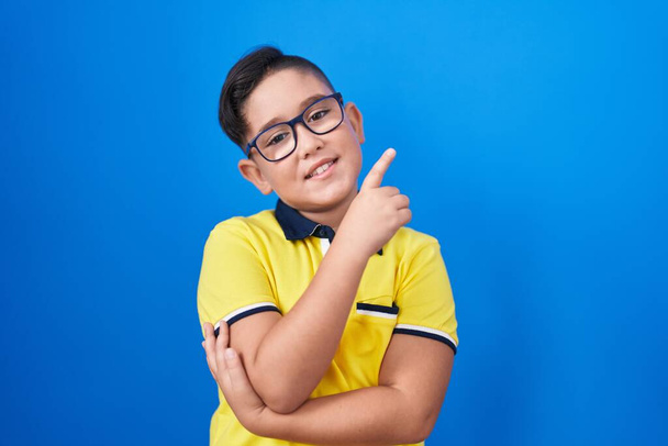 Молодий іспаномовний хлопчик стоїть над синім тлом усміхнений щасливий вказуючи з рукою і пальцем в бік  - Фото, зображення