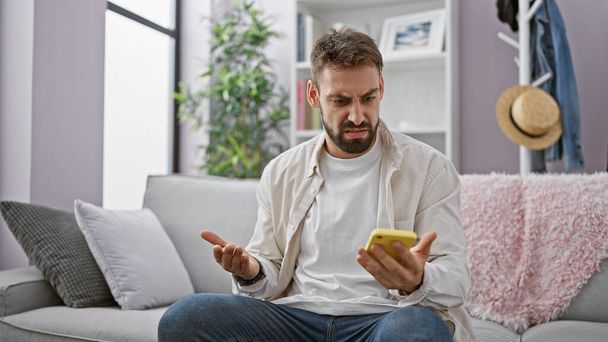Молодий, красивий іспаномовний чоловік з бородою сидить засмучений на дивані вдома, використовуючи смартфон. серйозний дорослий чоловічий смс, вдосконалений в технології в приміщенні. - Фото, зображення