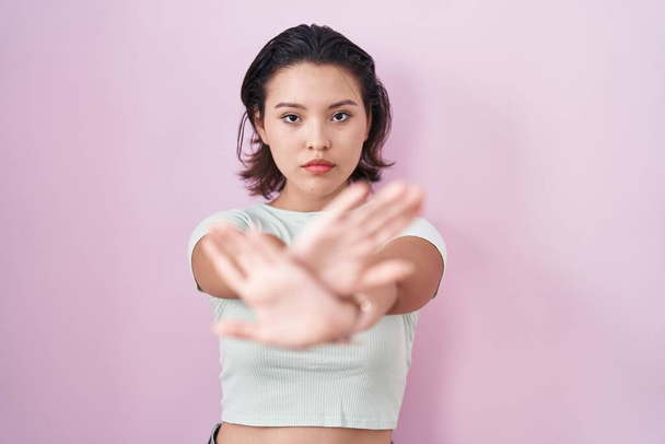 Latynoska młoda kobieta stojąca nad różowym tle odrzucenie wyrażenie skrzyżowanie ramion i dłoni robi negatywny znak, zły twarz  - Zdjęcie, obraz