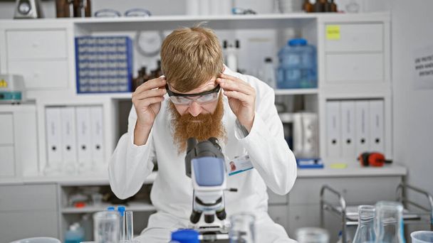 Guapo joven pelirrojo, un científico profesional, aprovechando el poder de la ciencia en un laboratorio, usando un microscopio y gafas en un trabajo de investigación serio. - Foto, Imagen