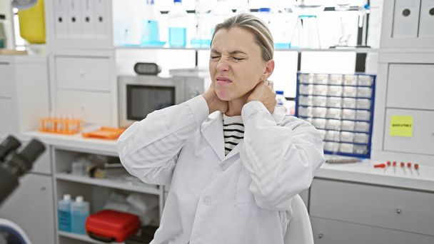 Una giovane donna caucasica con un camice da laboratorio ronza nel dolore, toccandosi il collo in un ambiente di laboratorio, ritraendo disagio o lesioni.. - Foto, immagini