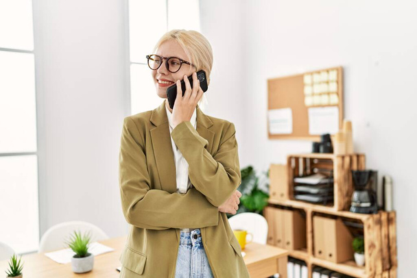 Νεαρή ξανθιά εργαζόμενη επιχειρηματίας που μιλάει στο smartphone και εργάζεται στο γραφείο - Φωτογραφία, εικόνα