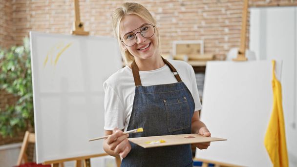 Jeune femme blonde artiste souriante confiante tenant pinceau et palette au studio d'art - Photo, image