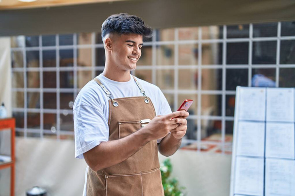 Νεαρός Ισπανός σερβιτόρος χαμογελώντας με αυτοπεποίθηση χρησιμοποιώντας smartphone στη βεράντα της καφετέριας - Φωτογραφία, εικόνα
