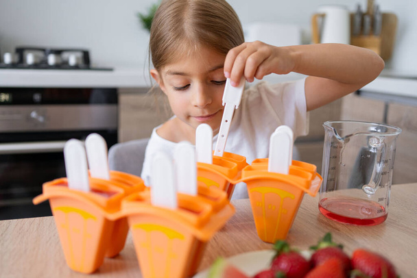 The process of making ice cream. Child making tasty ice lollipops in moulds on countertop in kitchen during free time. - Foto, imagen