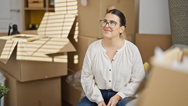 Молода красива іспаномовна жінка посміхається, дивлячись навколо сидячи на дивані в новому будинку - Фото, зображення