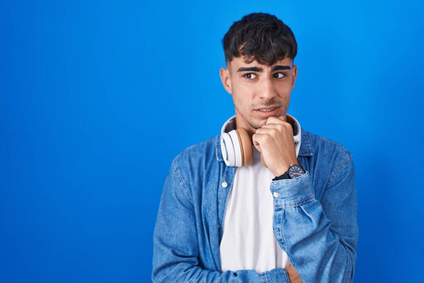 Giovane uomo ispanico in piedi sopra sfondo blu pensando preoccupato per una domanda, preoccupato e nervoso con mano sul mento  - Foto, immagini