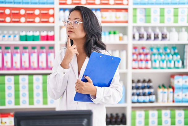Asiática joven mujer trabajando en farmacia celebración portapapeles cara seria pensando en la pregunta con la mano en la barbilla, reflexivo acerca de la idea confusa  - Foto, Imagen