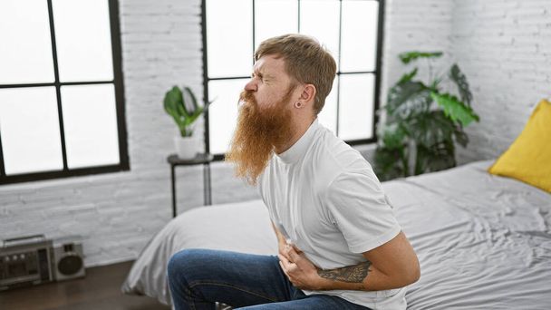 Tormented young adult redhead guy touching his aching stomach while sitting miserably on bed in bedroom, suffering from stomach pain, an indoor awakening ordeal - Photo, Image