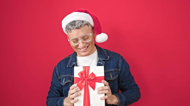 młody kaukaski mężczyzna uśmiechnięty pewny siebie trzymając świąteczny prezent nad odizolowanym czerwonym tle - Zdjęcie, obraz