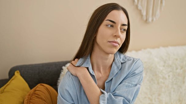 Beautiful young hispanic woman, sitting in cozy indoor living room, suffering intense back pain, stress etched on her face, doubting what to do next. - Photo, Image