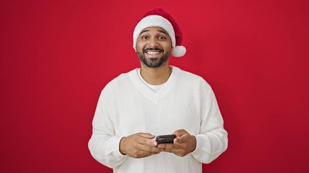Африканський американець у різдвяному капелюсі за допомогою смартфона над ізольованим червоним тлом - Фото, зображення