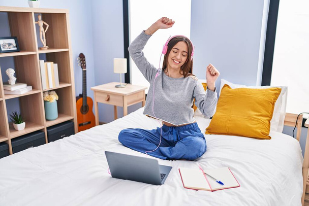 Young beautiful hispanic woman student listening to music studying on bed at bedroom - Photo, Image