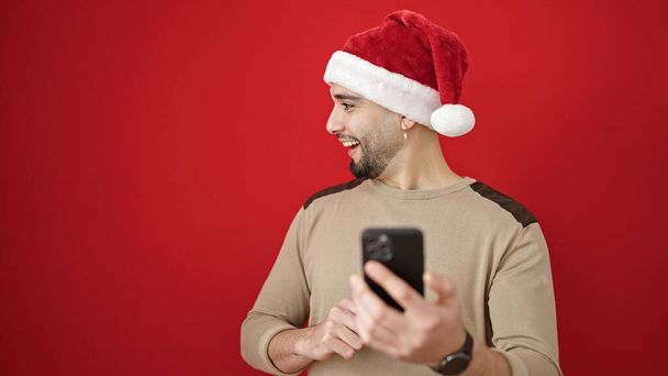 młody arabski człowiek noszący świąteczny kapelusz za pomocą smartfona na izolowanym czerwonym tle - Zdjęcie, obraz