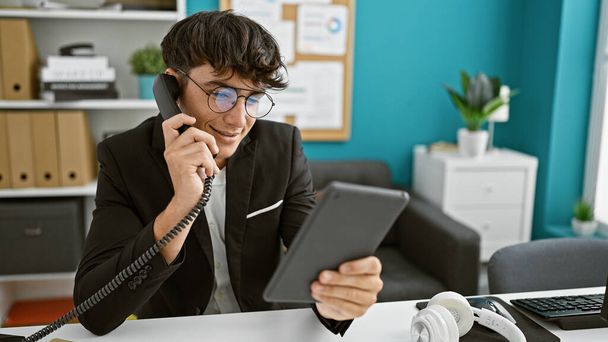 Charming young hispanic teenager triumphantly bossing his business talk over the phone at the office, scrolling touchpad with a sunny smile - Photo, Image