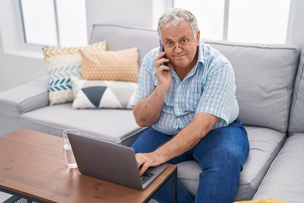 Седовласый мужчина средних лет разговаривает на смартфоне, используя ноутбук, сидя дома на диване. - Фото, изображение