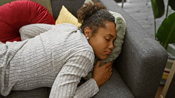 Uma jovem mulher com cabelo encaracolado dorme tranquilamente em um sofá cercado por almofadas em uma acolhedora sala de estar. - Foto, Imagem