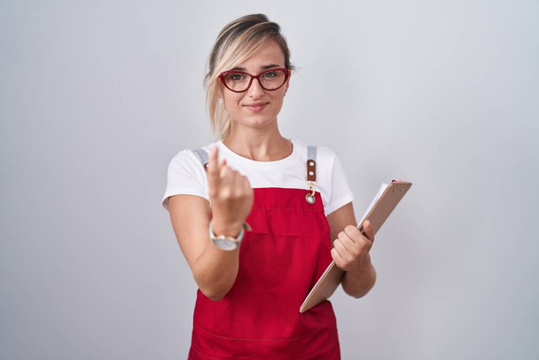Joven mujer rubia vistiendo uniforme de camarero sujetando portapapeles haciendo señas ven aquí gesto con la mano invitando a dar la bienvenida feliz y sonriente  - Foto, Imagen