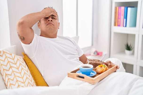 Jeune homme hispanique manger le petit déjeuner dans le lit stressé et frustré avec la main sur la tête, visage surpris et en colère  - Photo, image