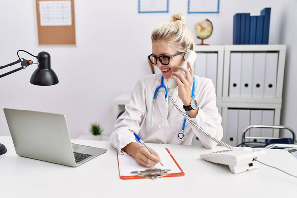 Νεαρή ξανθιά γυναίκα γιατρός μιλάει στο τηλέφωνο γράφοντας σε έγγραφο στην κλινική - Φωτογραφία, εικόνα
