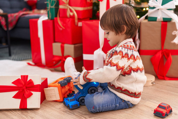 Entzückendes Kleinkind spielt mit LKW-Spielzeug, das zu Hause am Weihnachtsbaum auf dem Boden sitzt - Foto, Bild