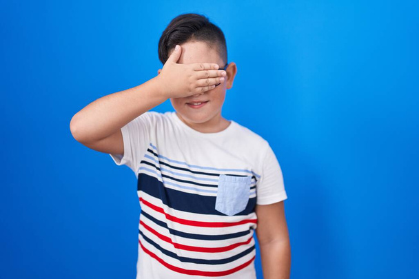 Ein junges hispanisches Kind, das vor blauem Hintergrund steht, lächelt und vor Überraschung mit der Hand auf dem Gesicht lacht. Blindes Konzept.  - Foto, Bild
