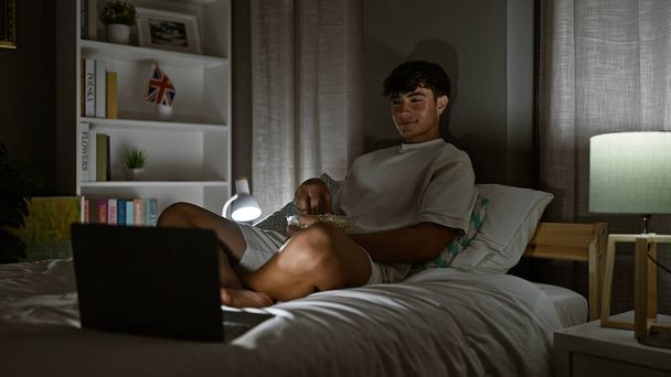 Atrapado joven adolescente hispano, cómodamente sentado en su cama, absorto en ver una película en su computadora portátil, irradiando un ambiente positivo en su acogedor dormitorio por la noche, haciendo que cada píxel cuente. - Foto, Imagen