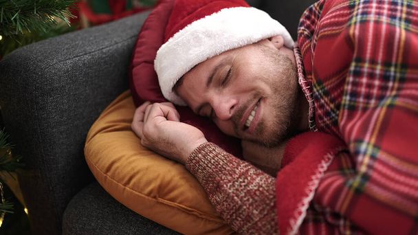 Молодой кавказский мужчина лежит на диване и спит в рождественской шляпе дома. - Фото, изображение
