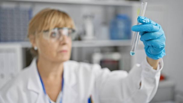 Científica rubia de mediana edad, vestida con gafas y guantes, mide meticulosamente el contenido del tubo de ensayo en un laboratorio bullicioso. - Foto, Imagen