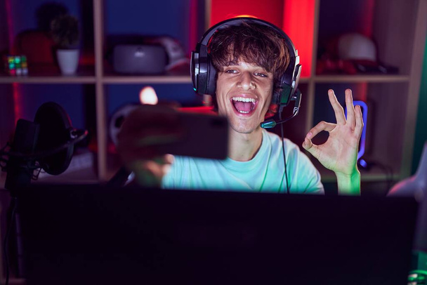 Latino nuori mies pelaa videopelejä älypuhelin tekee ok merkki sormin, hymyillen ystävällinen ele erinomainen symboli  - Valokuva, kuva