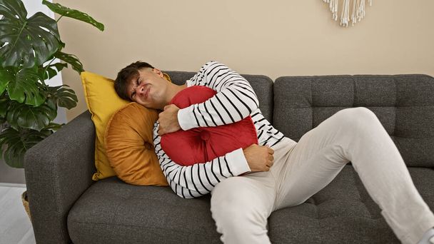 Занепокоєний молодий іспаномовний чоловік страждає від болю в животі, нещадно лежачи на домашньому дивані, обіймаючи подушку, в інтер'єрі кімнати, що вказує на серйозну хворобу - Фото, зображення