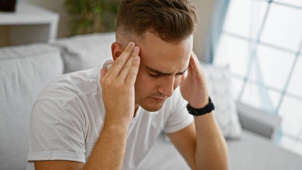 Молодой взрослый мужчина, выглядящий напряженным в помещении, возможно испытывающий головную боль или стресс, на фоне нейтральной спальни. - Фото, изображение