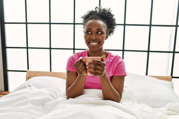 African american woman drinking cup of coffee sitting on bed at bedroom - Fotoğraf, Görsel