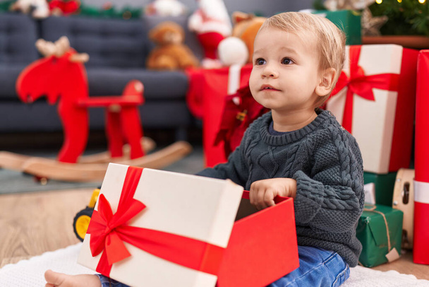Uroczy blond maluch rozpakowywanie prezent świąteczny siedzi na podłodze w domu - Zdjęcie, obraz