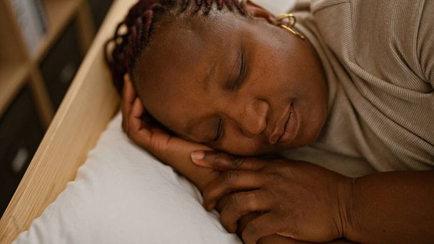 Esausta donna afroamericana che trova conforto rilassandosi nella sua accogliente camera da letto, sdraiata addormentata sul suo comodo letto, riposata stanca sotto una morbida coperta, capelli intrecciati sparsi sul cuscino - Foto, immagini